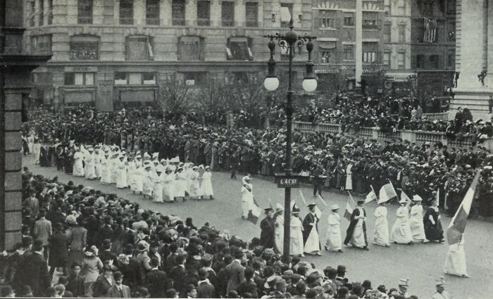 Women in the 20th century suffrage era marching in white dresses during a large public demonstration with crowds watching.