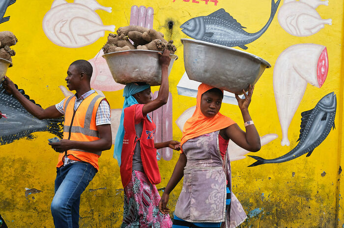 Three street vendors carrying goods on their heads walk past a vibrant yellow mural of fish and meat in a busy urban scene.