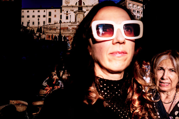 Close-up of a woman wearing large sunglasses with another woman and historic buildings in the background in a street photo.