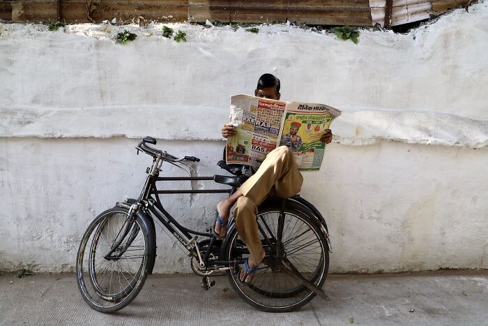 Man sitting on a bicycle against a white wall, reading a newspaper in a street photo capturing everyday life art.