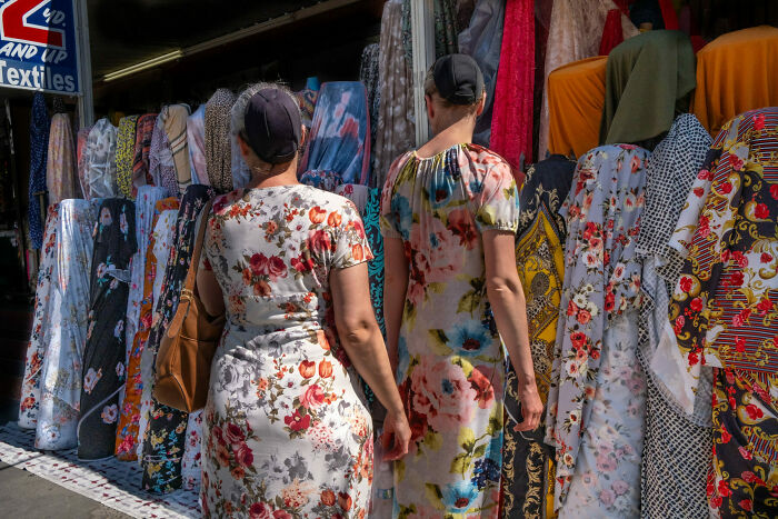 Two women in floral dresses walk past colorful fabric rolls at a street market, capturing unforgettable street photos.