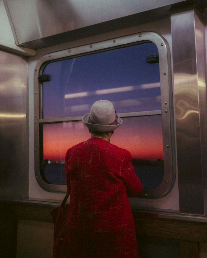 Woman in a red coat and hat looking out a window at a vibrant sunset, a striking example of street photography art.