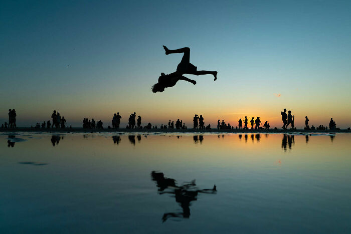 Silhouetted person flipping midair above water reflecting sunset, surrounded by crowds in unforgettable street photos.