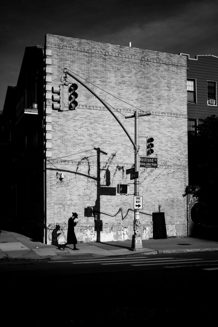 Black and white street photo of a person walking at a city corner with traffic lights and urban architecture shadows.