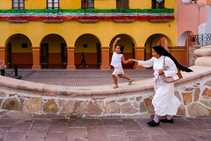Child joyfully balancing on a wall while holding hands with a nun in colorful street, captured in unforgettable street photos.