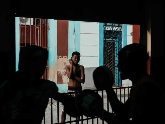 Silhouettes of two people boxing with gloves while a boy stands in the background in a colorful street setting, street photos.