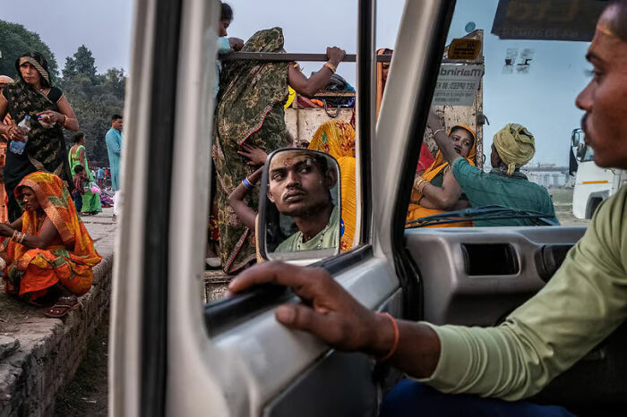 Man reflected in a side mirror of a vehicle with vibrant crowd of people in traditional clothing outside in a street photo.