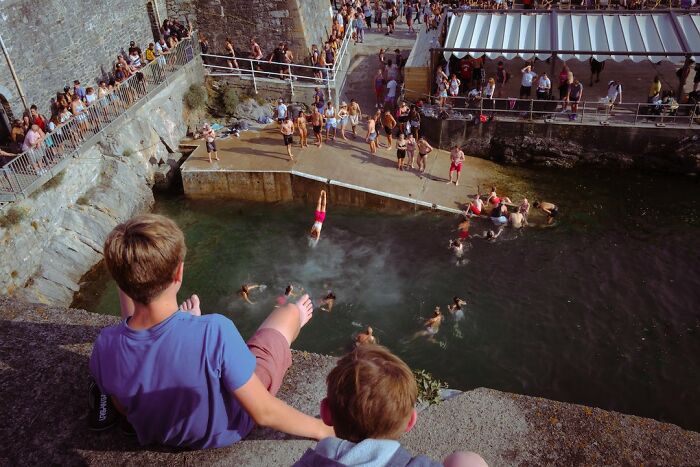 Two boys sitting on a ledge watching people swimming and diving in a natural water pool surrounded by stone walls.
