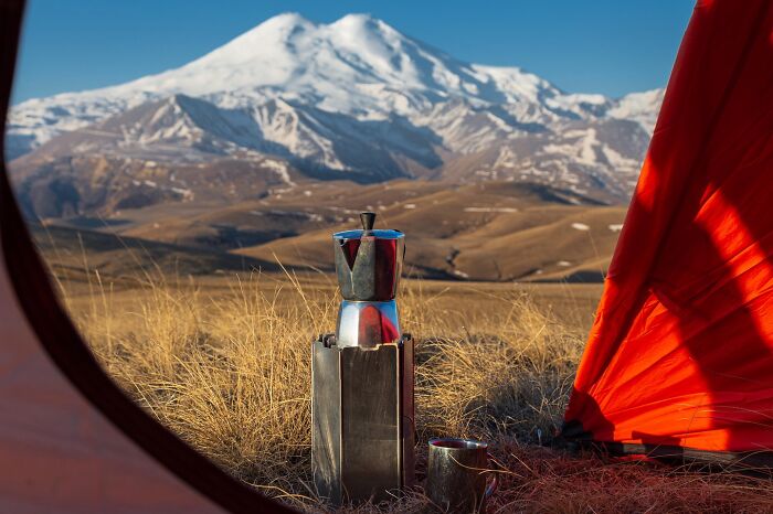 Camping scene showing a coffee pot and cup with stunning nature views of snow-capped mountains and dry grasslands.