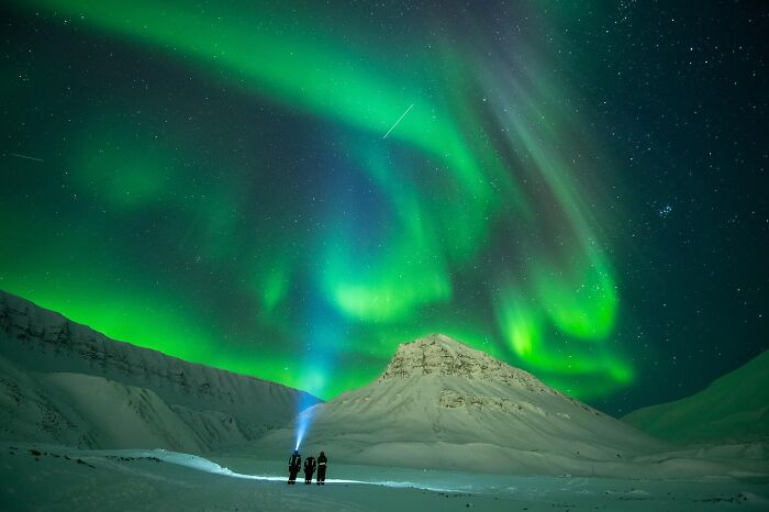 Three people stand on snowy terrain under stunning northern lights, capturing nature and life around the world.