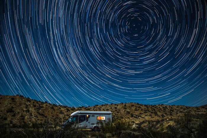 RV parked in desert under stunning star trails, capturing nature and life around the world at night.