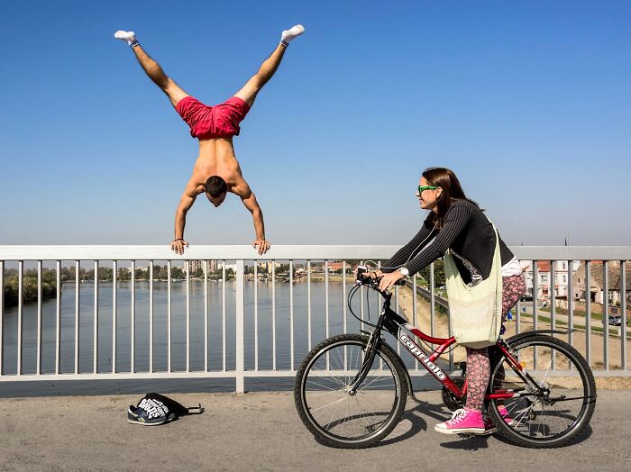Man doing handstand on railing near river while woman on bicycle watches in vibrant outdoor scene capturing nature and people.