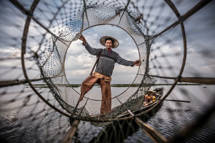 Fisherman balancing on boat using fishing net on calm water, capturing nature, people, and life around the world.