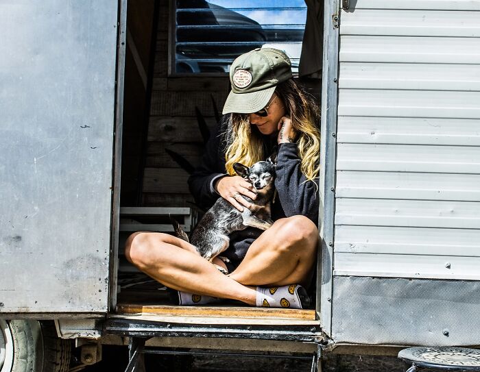 Woman wearing a cap sitting in a trailer doorway, holding a small dog, capturing a moment of life around the world.