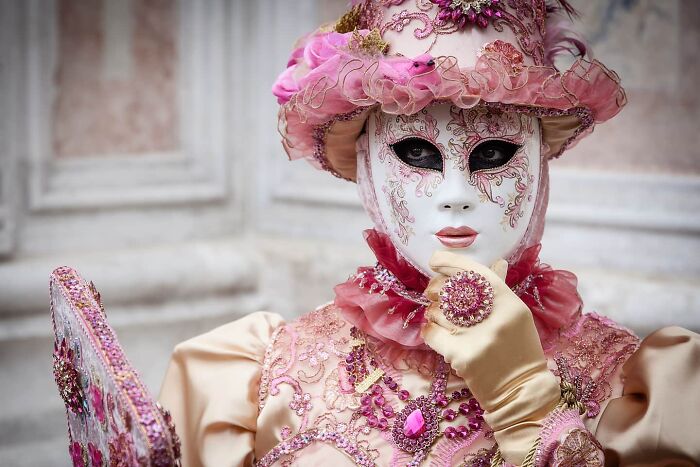 Person wearing elaborate Venetian mask and costume adorned with pink and purple details, capturing stunning people and life.