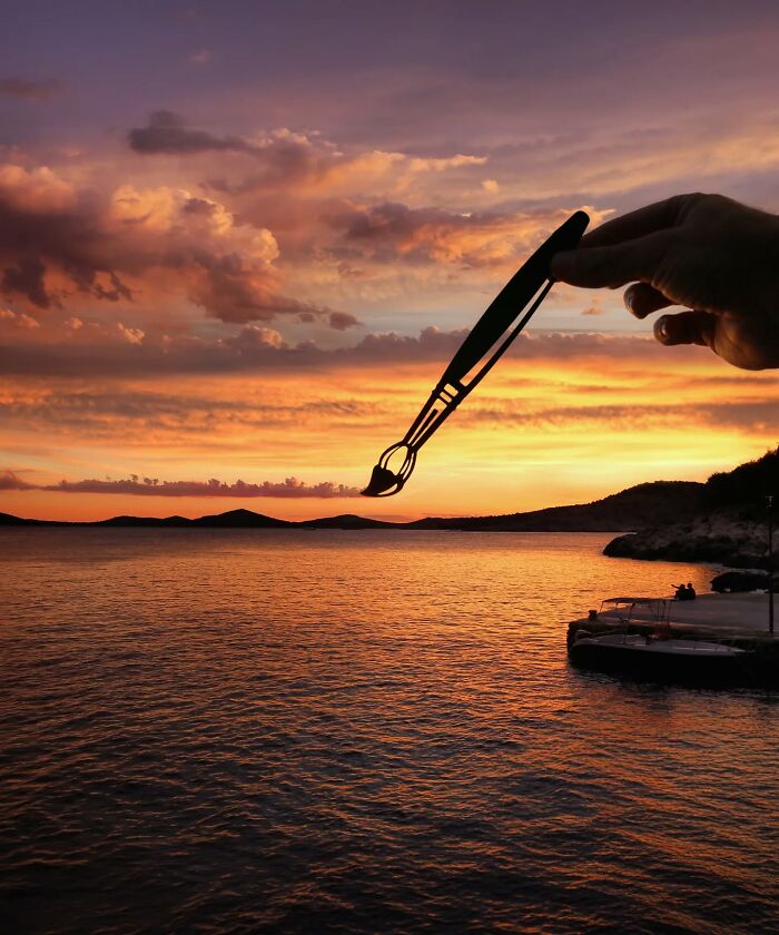Hand holding paper cutout of a paintbrush over a sunset sea scene, showcasing creative artist travel concept.