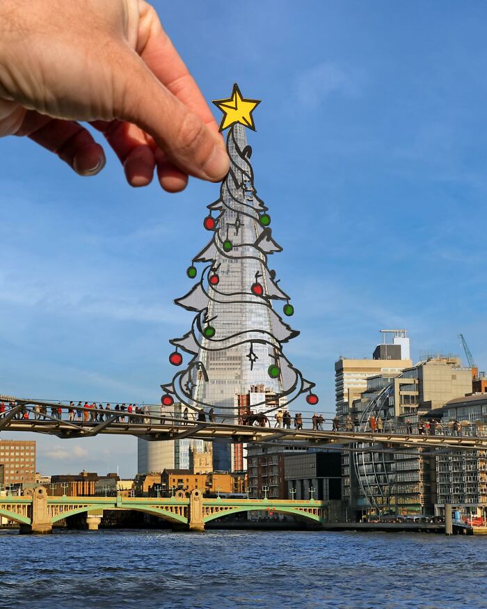 Hand holding paper cutout of a decorated Christmas tree over city skyline, showcasing artist traveling the world with paper cutouts.