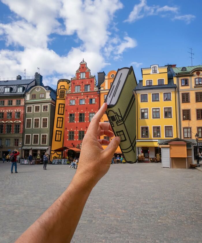 Hand holding a paper cutout book in front of colorful historic buildings, showcasing artist travels and creative scenes.