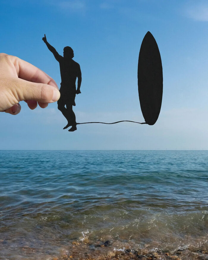 Hand holding paper cutouts of a surfer and surfboard creating a playful scene at the beach, showcasing paper cutouts art.