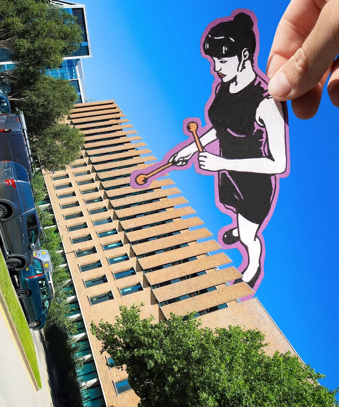 Hand holding a paper cutout figure playing xylophone on the side of a tall building under clear blue sky.