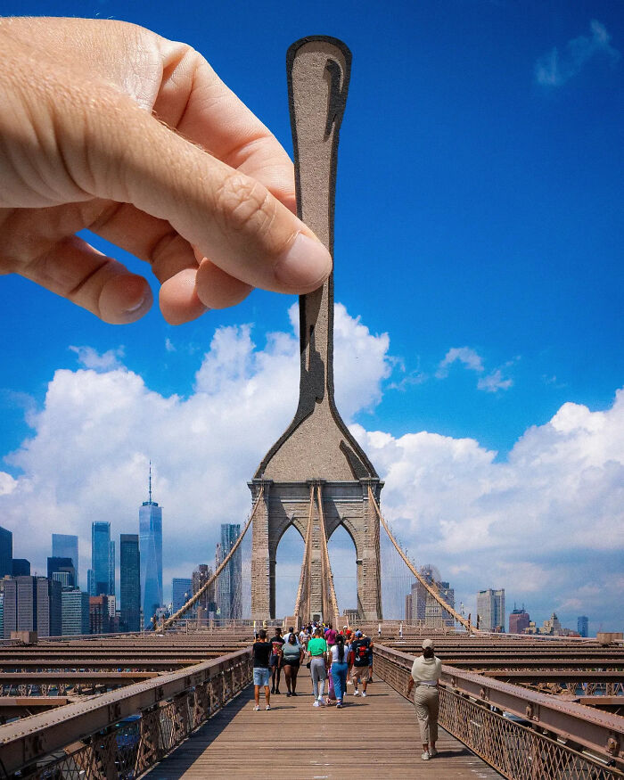 Hand holding paper cutout of a spoon aligned with Brooklyn Bridge, artist travels the world creating surprising scenes.