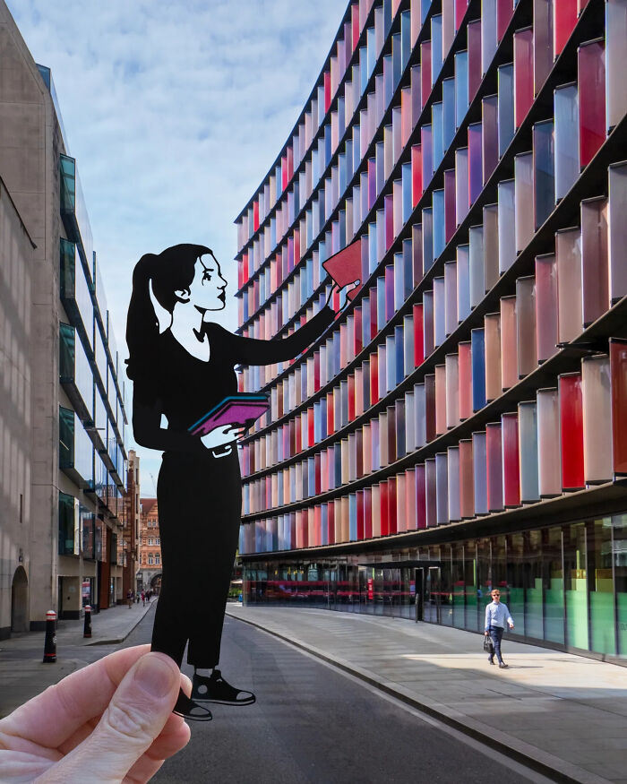 Hand holding paper cutout of a woman interacting with colorful building windows, artist travels the world with paper cutouts.