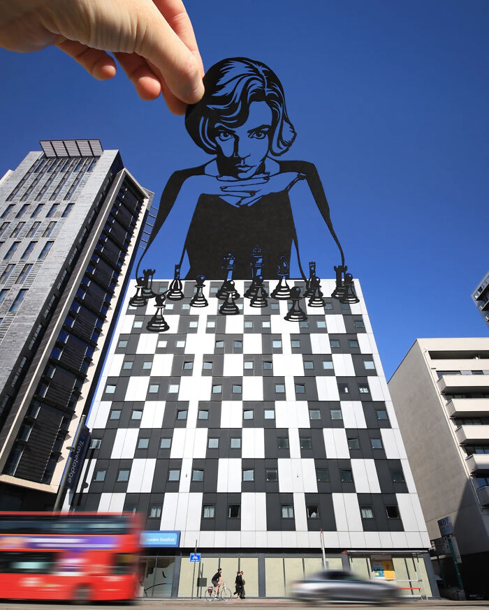 Hand holding paper cutout of a woman playing chess integrated with a building facade in artist travels the world scenes.