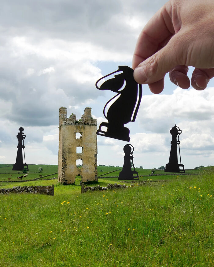 Hand holding black paper cutout chess knight among oversized cutouts in a green field, artist travels the world with paper cutouts.