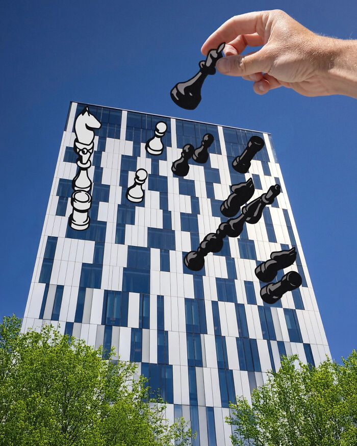 Hand holding paper cutout chess piece over building with windows arranged like a chessboard in artist's creative scene.