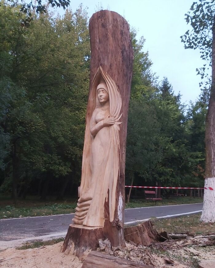 Chainsaw artist carves a detailed wooden sculpture of a woman from a forgotten tree trunk in an outdoor park setting.
