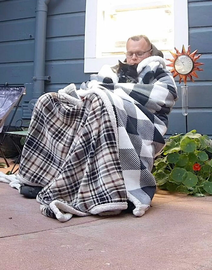 Man wrapped in blankets holding a tuxedo cat on a porch near a blue house and garden with a decorative thermometer. Man wrapped in blankets holding a tuxedo cat on a porch near a blue house and garden with a decorative thermometer.