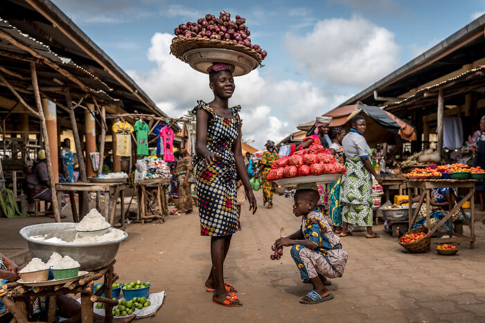 Woman and child carrying baskets of produce on their heads in a bustling market, showcasing the beauty of cultures.
