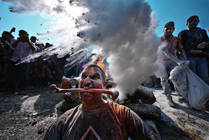 Man with red powder on face holding stick in mouth during vibrant cultural festival in India, bursting with color and life.
