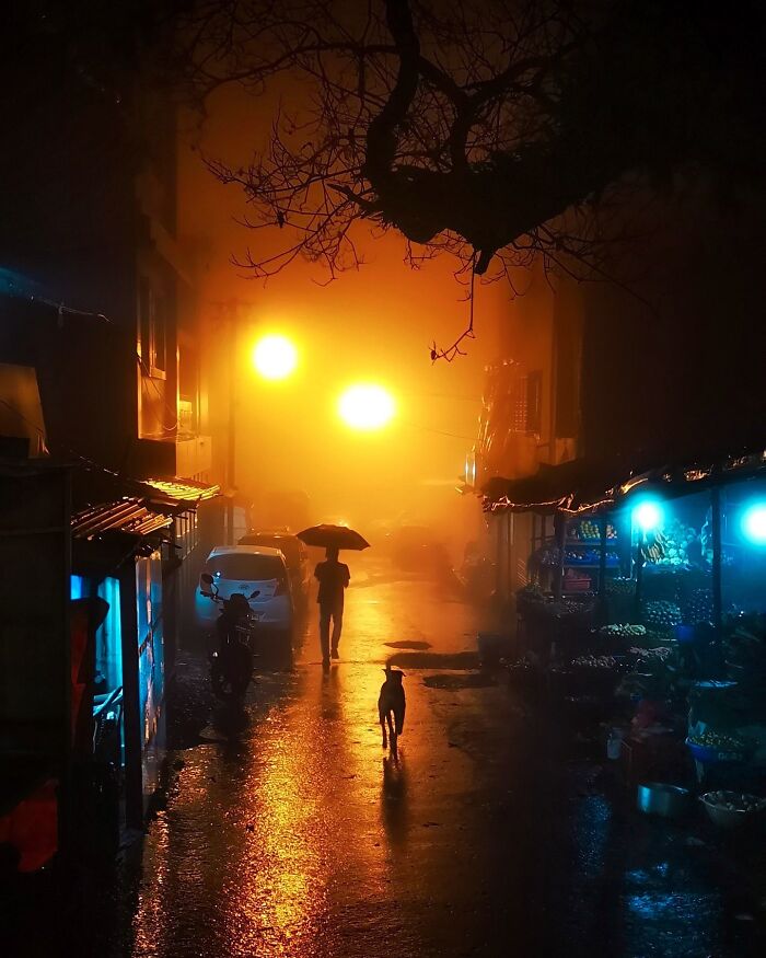 Person walking with umbrella on a wet street at night in India, vivid colors and life with glowing yellow lights and market stalls.
