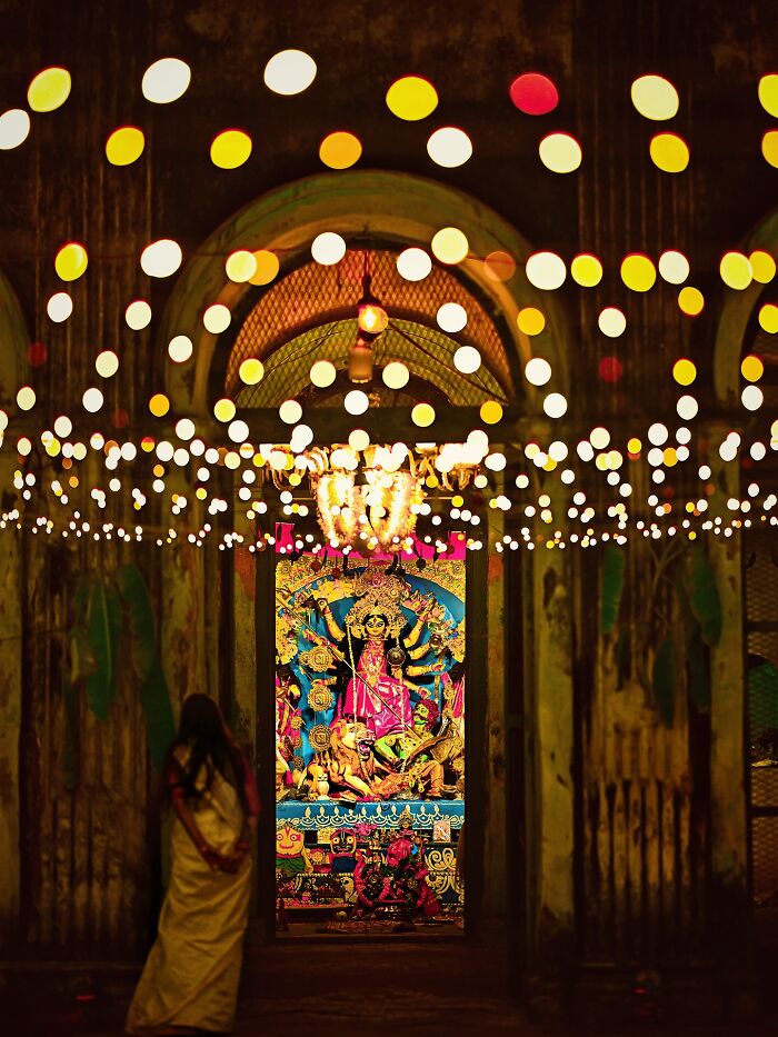 Vibrant photo of colorful Indian festival lights and a woman in a white saree admiring a traditional deity statue indoors.