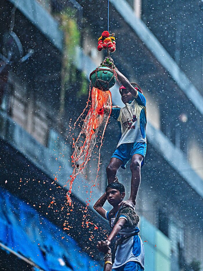 Two boys participating in a vibrant festival in India, breaking a decorated pot spilling orange liquid in rain.
