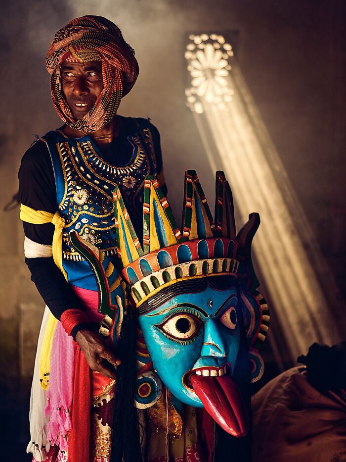 Man in traditional colorful attire holding a vibrant Indian mask with a blue face and red tongue indoors with light beams.