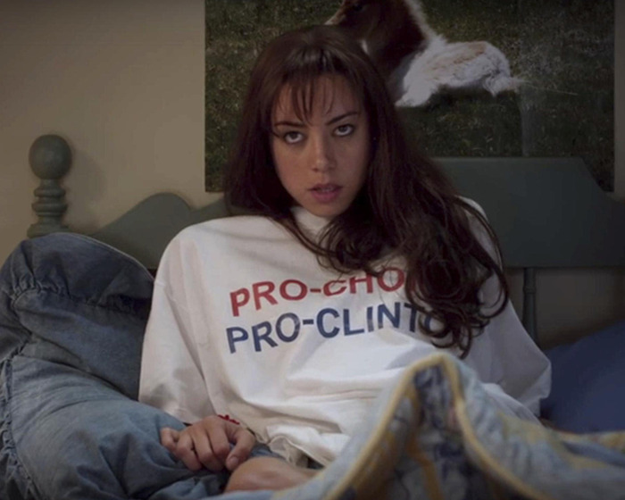 Young woman with long dark hair wearing a Pro-Choice Pro-Clinton shirt, sitting on a bed with casual expression.