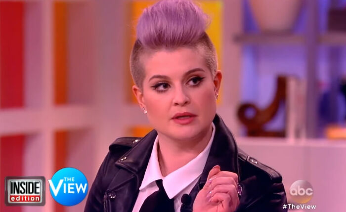 Celebrity with purple hair and leather jacket on a talk show discussing moments when celebs seemed out-of-touch trying to be relatable.