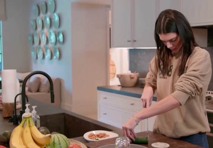 Celebrity in casual home kitchen setting, slicing cucumber, illustrating moments of celebs trying to be relatable.