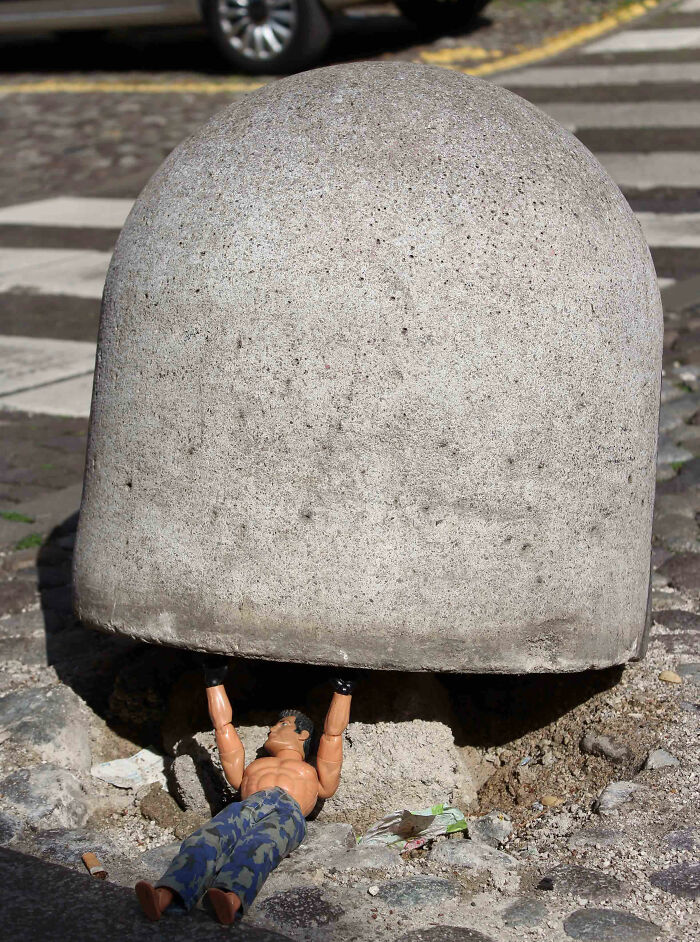 Street art photo showing a toy figure lifting a large concrete bollard on a city street canvas.