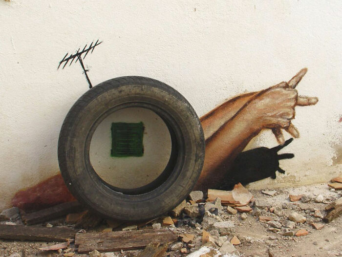 Street art photo of hands creating shadow puppets beside a tire and antenna against a white wall on rubble ground.