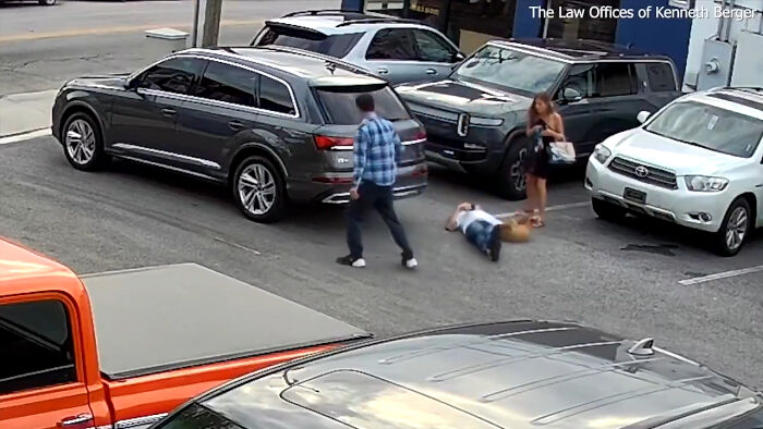 Man lying on ground near service dog after altercation with Uber driver in parking lot, woman standing nearby watching.