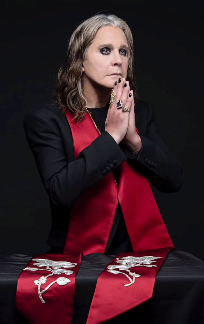 Rock legend with long hair and dark eye makeup, dressed in black and red, posing thoughtfully for rock legends gathering.