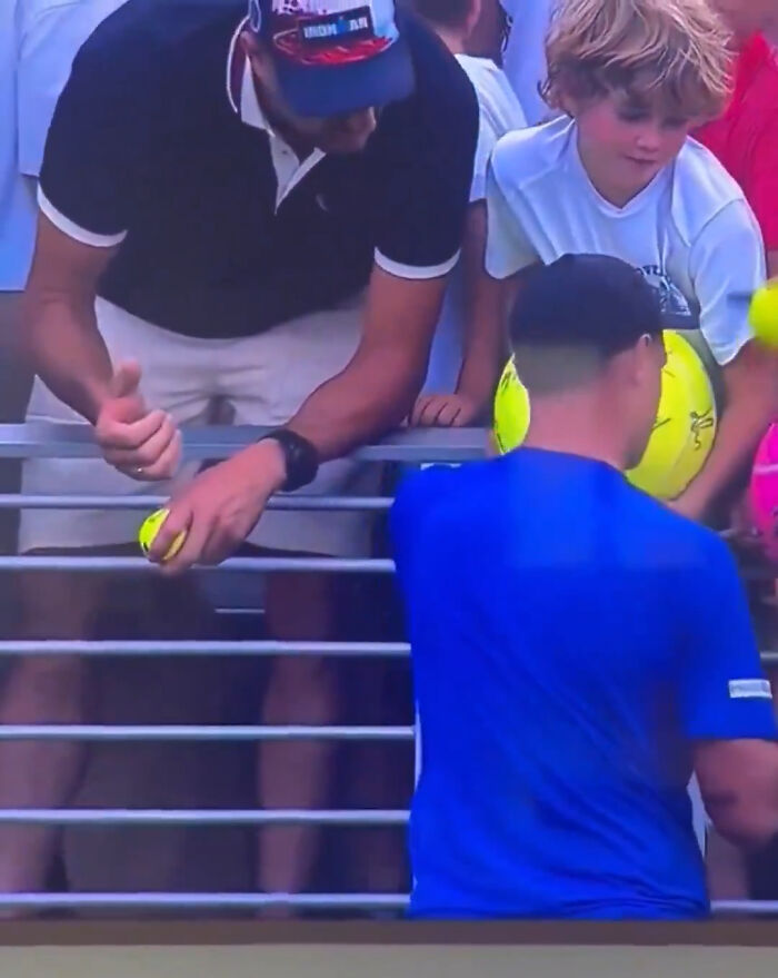 Polish CEO caught at US Open reaching for a fan's hat, surrounded by tennis balls and spectators.