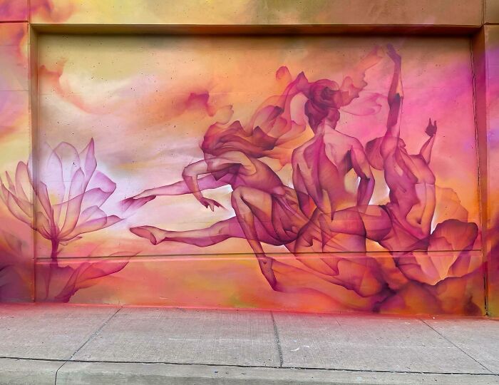 Abstract mural blending graffiti and realism depicting flowing female figures and a large flower in warm pink and orange tones.