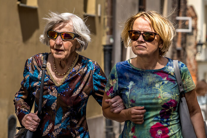 Two women wearing sunglasses walking arm in arm on a sunny street, showcasing the beauty of cultures captured by a photographer.