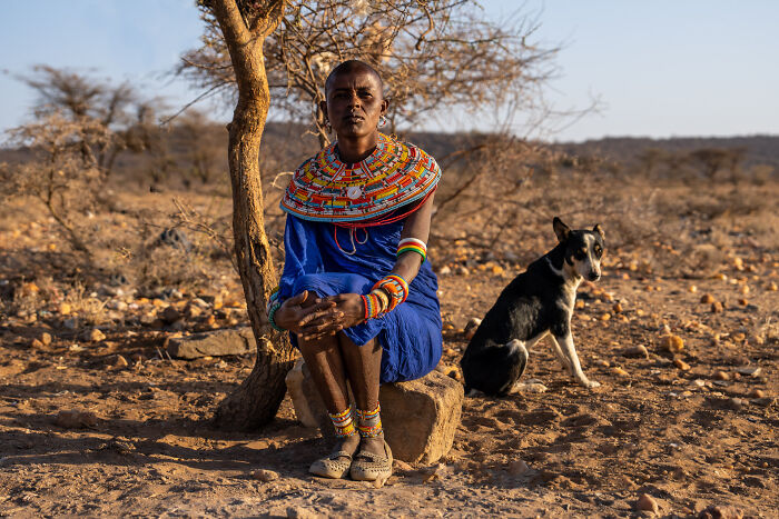 Masai woman in traditional vibrant attire sitting by a tree in a rural landscape, showcasing cultural beauty from travels.
