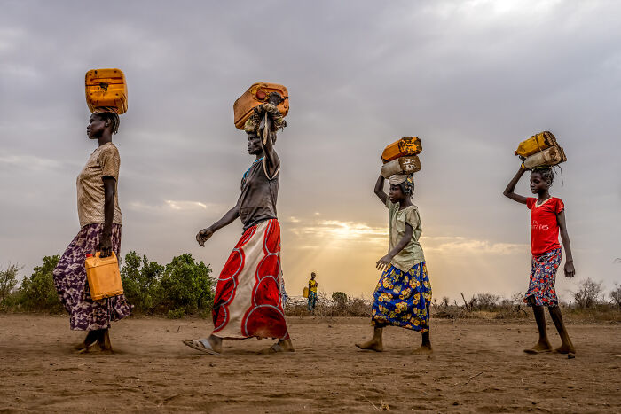 Women carrying yellow containers on their heads in a rural landscape, showcasing the beauty of cultures through travel photography.