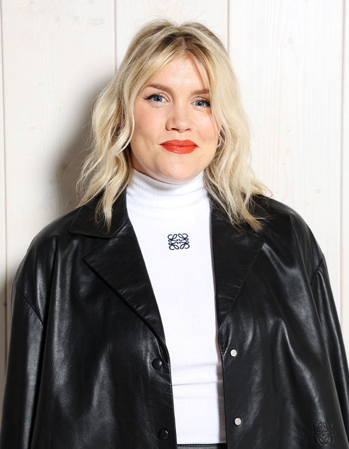 Blonde woman wearing black leather jacket and white turtleneck, posing against light wooden wall for Wuthering Heights adaptation.